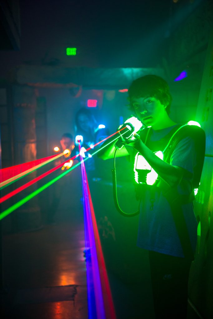 Jugendlicher spielt Lasertag mit leuchtender Sensorweste und Markierer, während farbige Laserstrahlen durch die dunkle Arena führen.