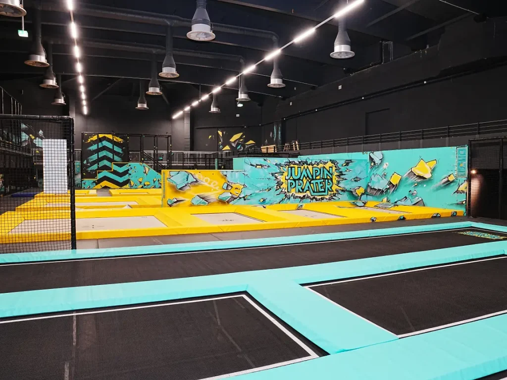 Leere Trampolinhalle mit schwarzen Sprungflächen, türkisfarbenen Rahmen und gelbem Bereich vor einer bunt gestalteten Wand.