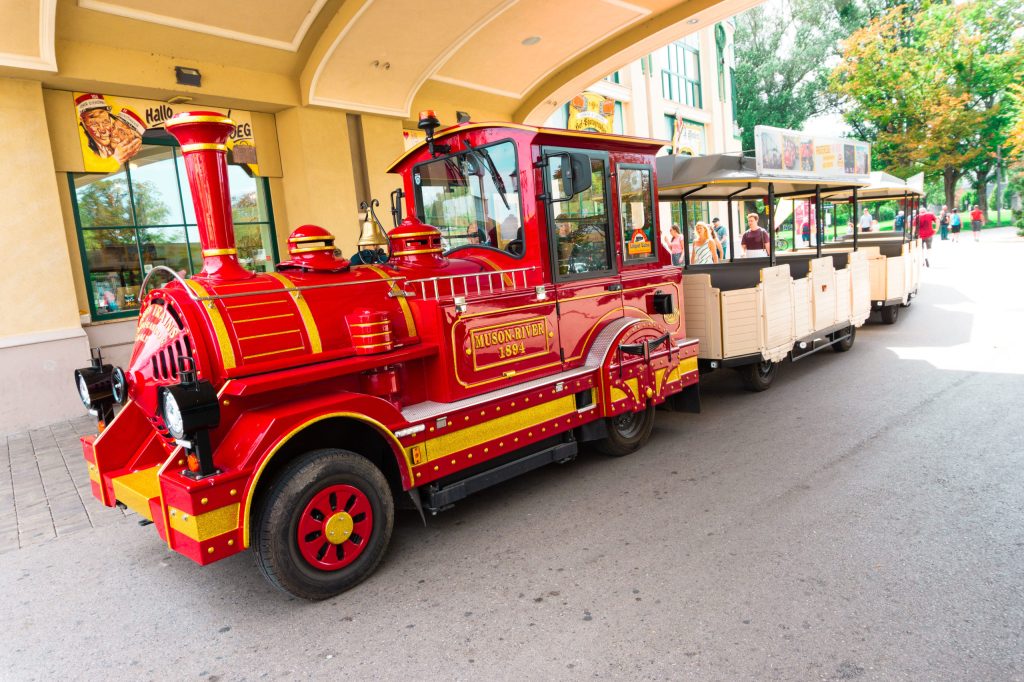 Rote Parkbahn im Stil einer Dampflokomotive mit mehreren offenen Personenwagen vor einem Gebäude.