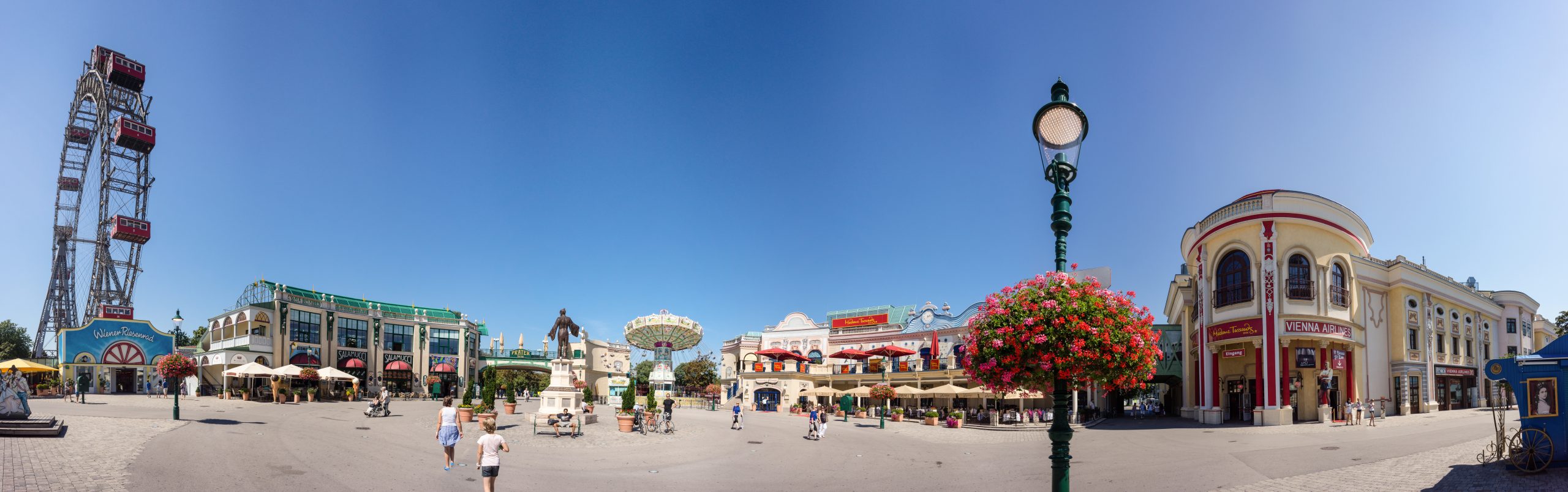 Panoramaaufnahme des Wiener Praters mit dem Riesenrad, dem Madame Tussauds Gebäude und umliegenden Attraktionen bei strahlendem Sonnenschein