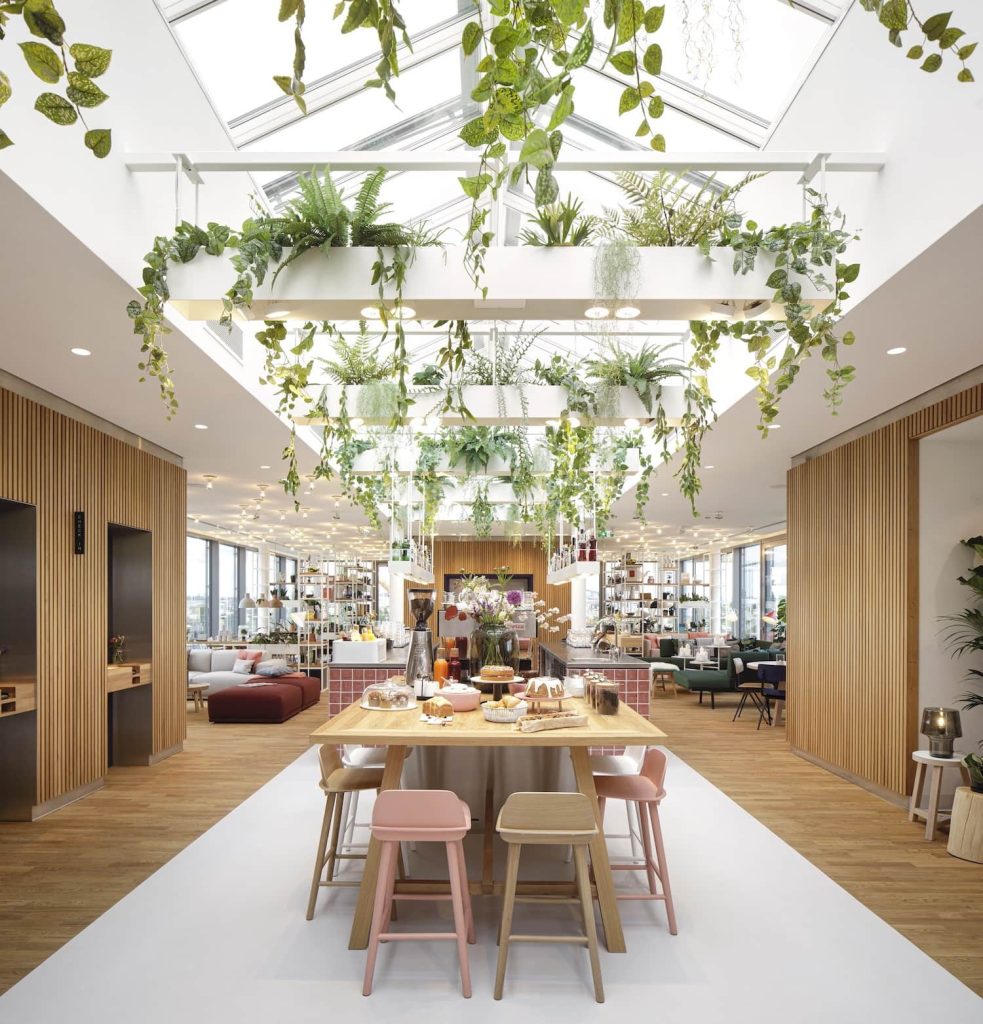 Heller Lounge- und Essbereich mit Holztisch, Sitzgruppen, großen Dachfenstern und üppig hängenden Grünpflanzen.