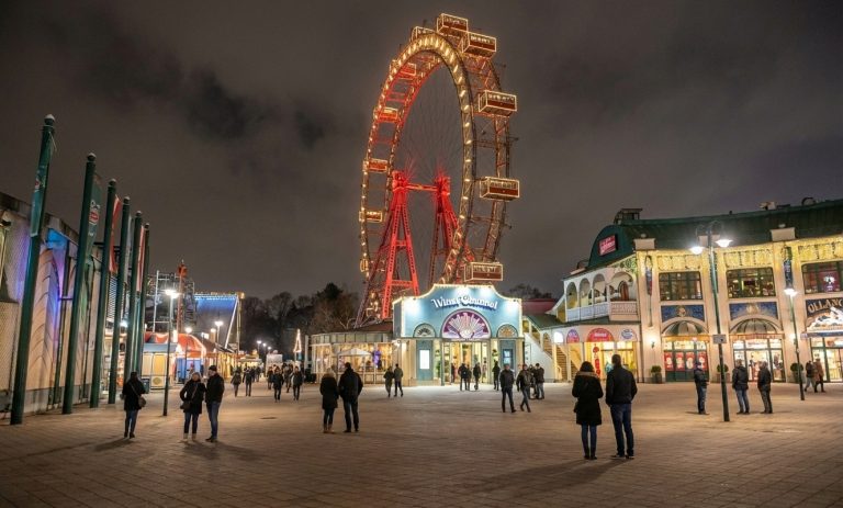 Wiener Riesenrad bei Nacht im Prater, festlich beleuchtet über dem Eingangsbereich und umgeben von Besuchern auf dem Platz.