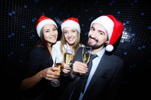 Drei Personen mit Weihnachtsmützen stoßen bei einer Weihnachtsfeier mit Sektgläsern an und lächeln in die Kamera.
