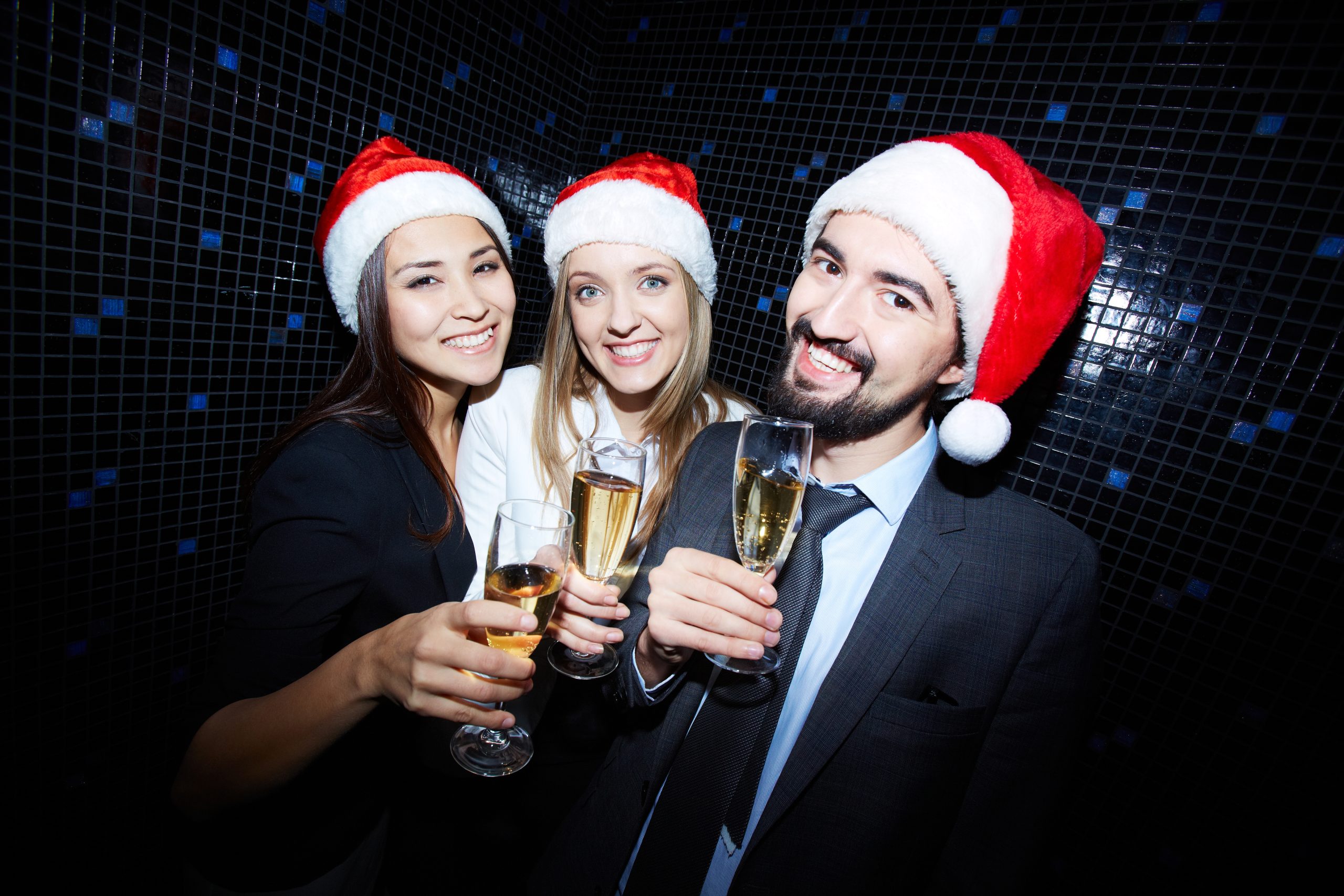 Drei Personen mit Weihnachtsmützen stoßen bei einer Weihnachtsfeier mit Sektgläsern an und lächeln in die Kamera.