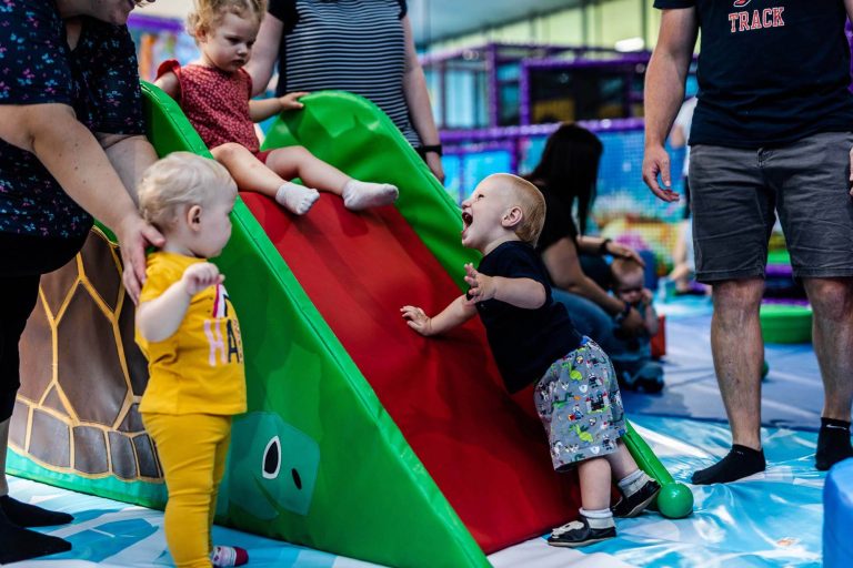 Kleinkinder spielen an einer bunten Indoor-Rutsche in einer weichen Spielhalle mit Erwachsenen im Hintergrund