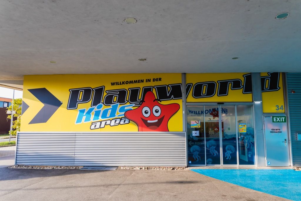 Eingang der Playworld Kids Area mit gelbem Logo, rotem Stern-Maskottchen und Eingangstüren einer Indoor-Spielhalle