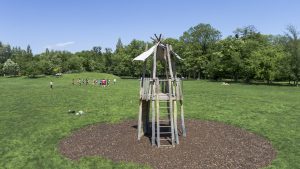 Holz-Spielturm mit Leiter auf einer großen Wiese in einem sonnigen Park mit Bäumen im Hintergrund