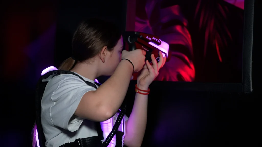 Mädchen mit Lasertag-Weste und Phaser in Deckung, konzentriert beim Zielen in einer dunklen Indoor-Lasertag-Arena.