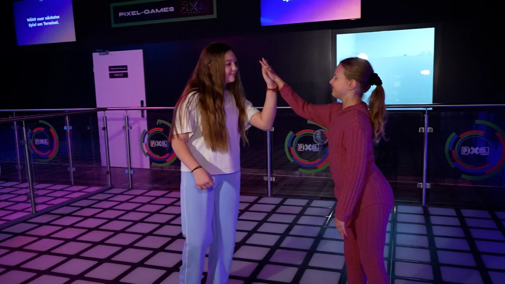 Zwei Kinder geben sich ein High-Five auf einer beleuchteten Tanzfläche in einer modernen Indoor-Freizeitanlage.